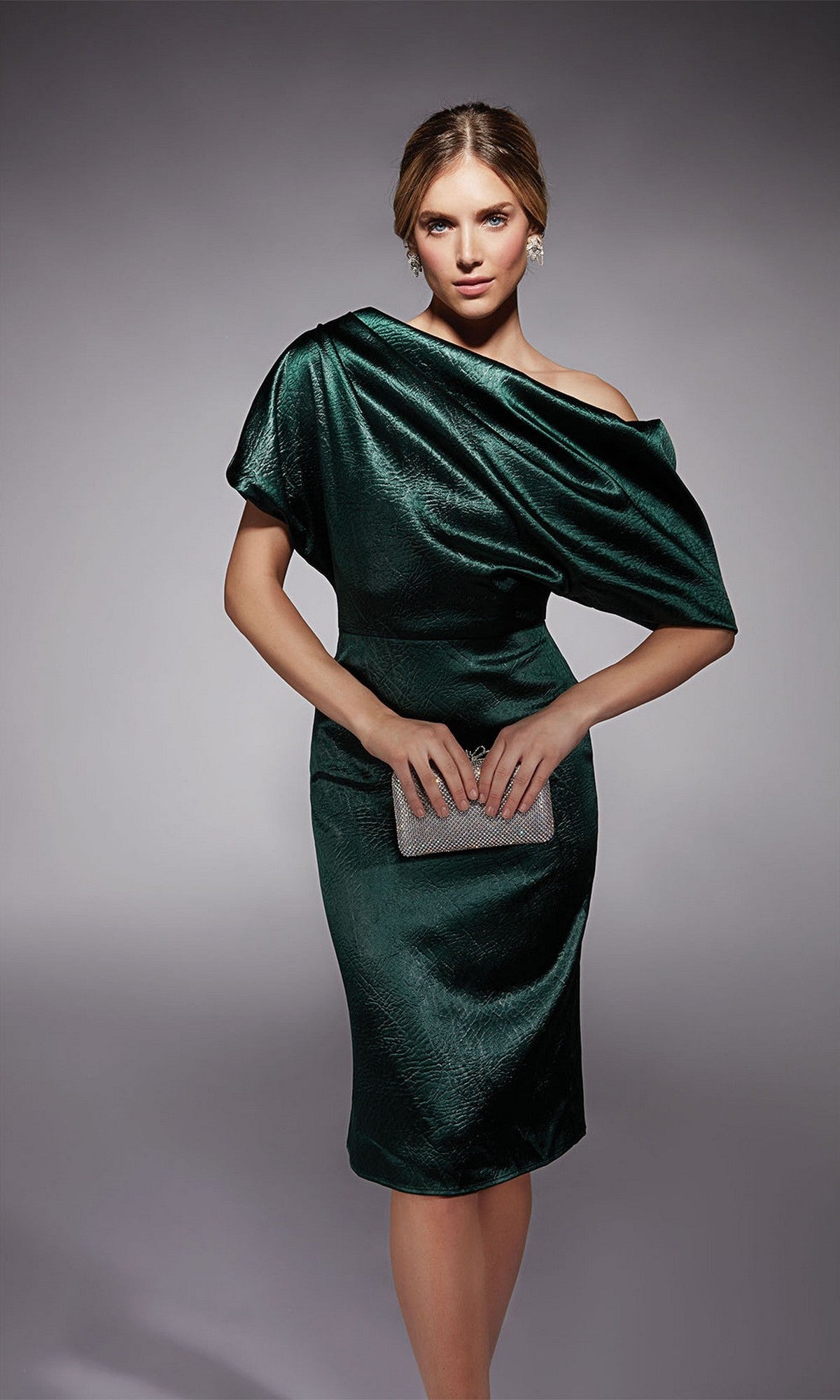 Woman wearing the Alyce 70060 knee length semi-formal dress in forest green, holding a silver clutch against a plain gray background.